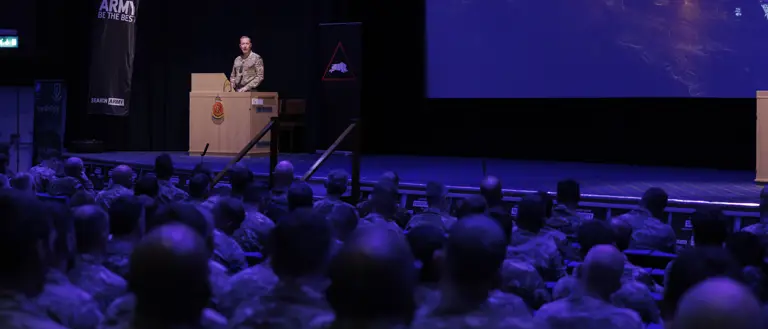 A military officer stands at a podium addressing uniformed soldiers seated in an auditorium. A screen reads