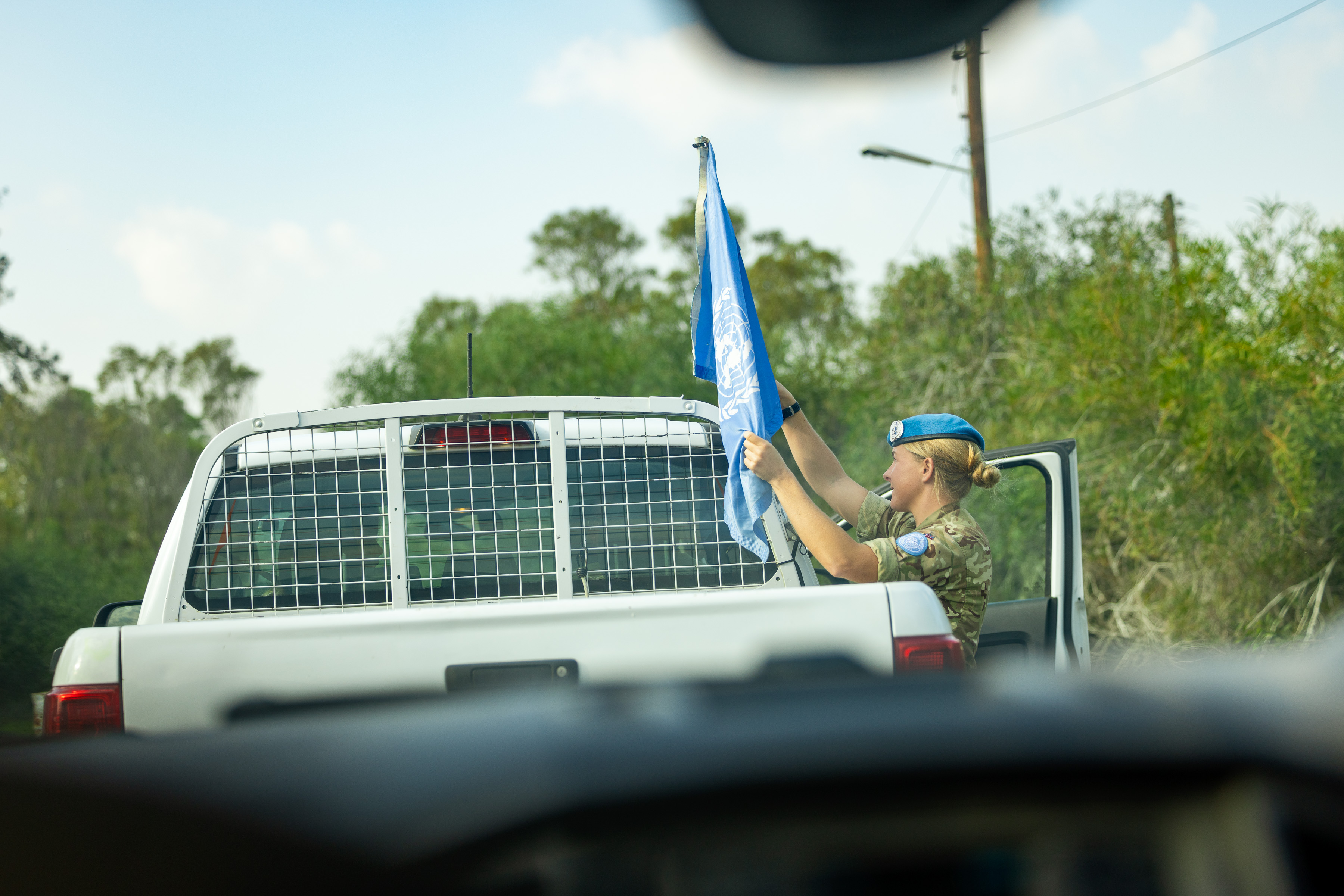 A woman wearing uniform and a blue beret is picture adjusting a United Nations flag atop a car. 