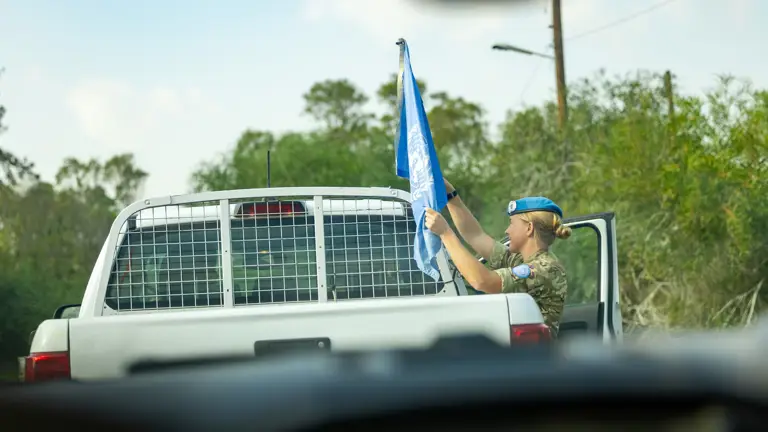 A woman wearing uniform and a blue beret is picture adjusting a United Nations flag atop a car.