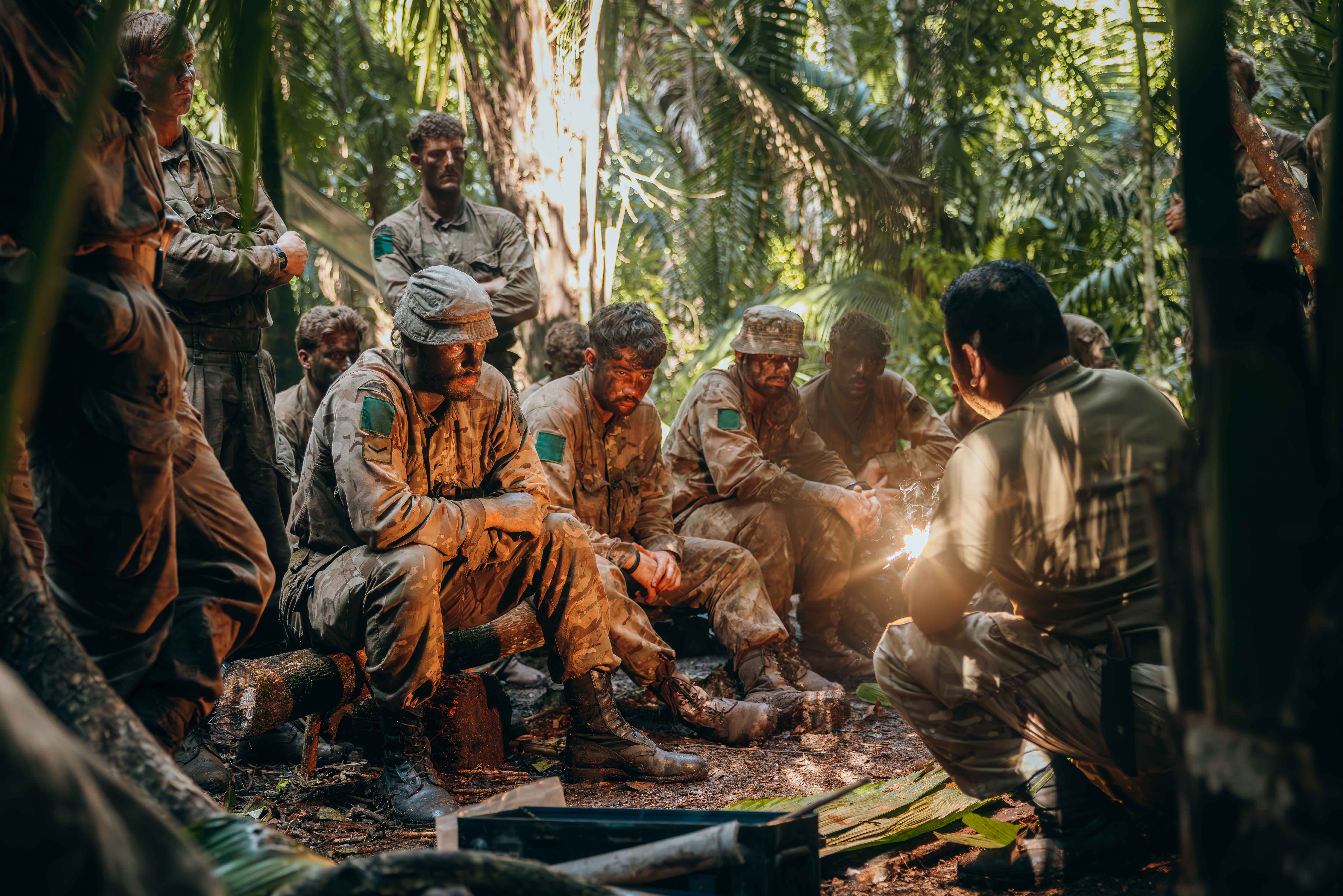 Group of soldiers in camouflage uniforms sitting and listening attentively to a briefing in a dense jungle setting.
