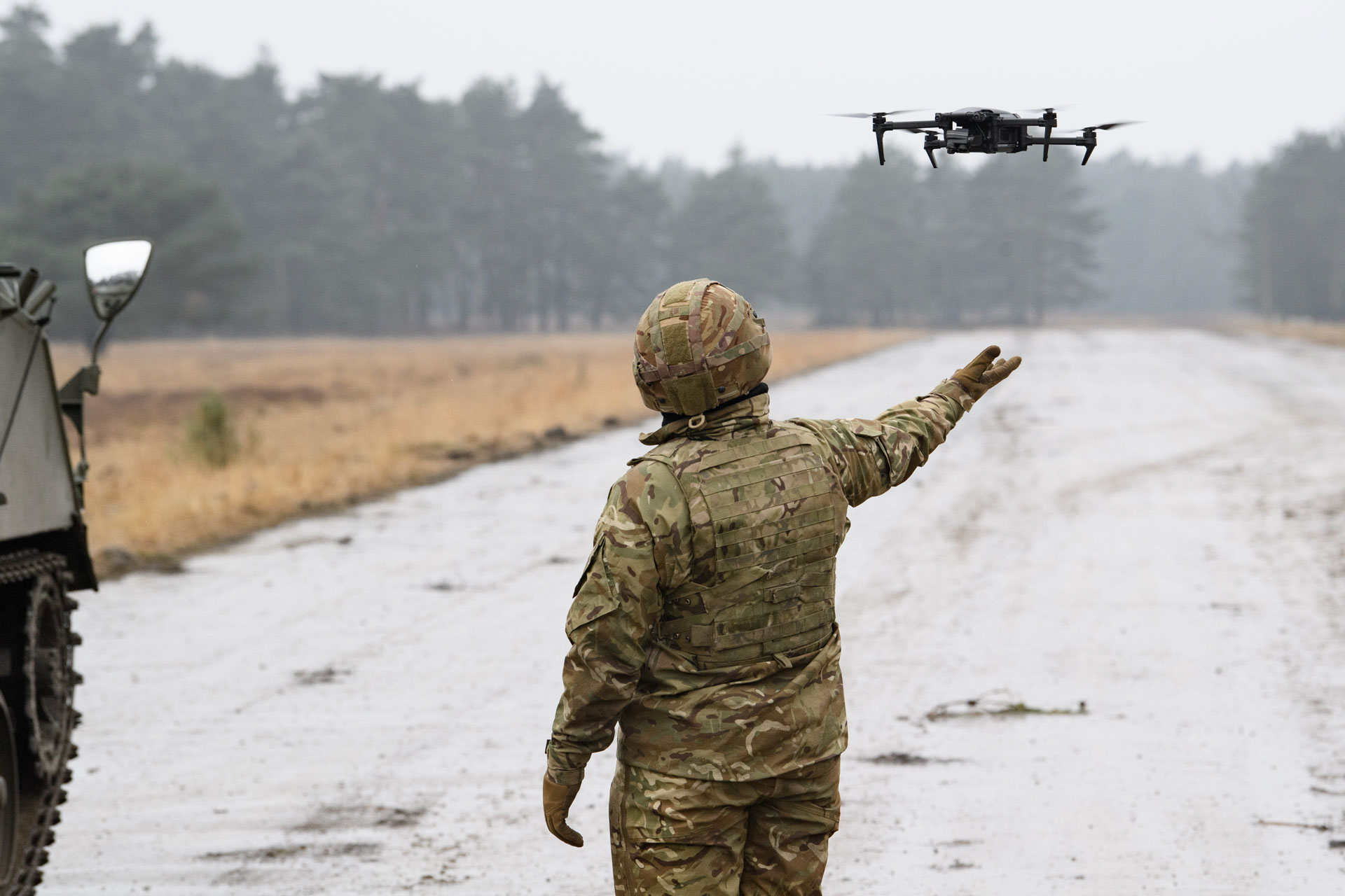 A soldier in camouflage uniform controls a drone hovering at arm's length on a rural road next to a military vehicle.