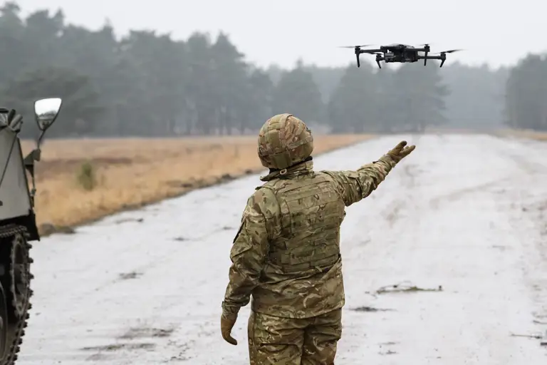 A soldier in camouflage uniform controls a drone hovering at arm's length on a rural road next to a military vehicle.