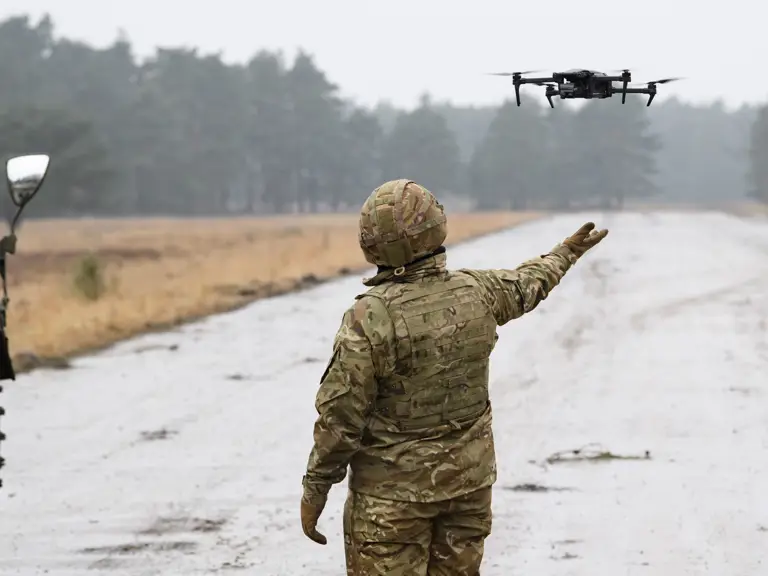 A soldier in camouflage uniform controls a drone hovering at arm's length on a rural road next to a military vehicle.