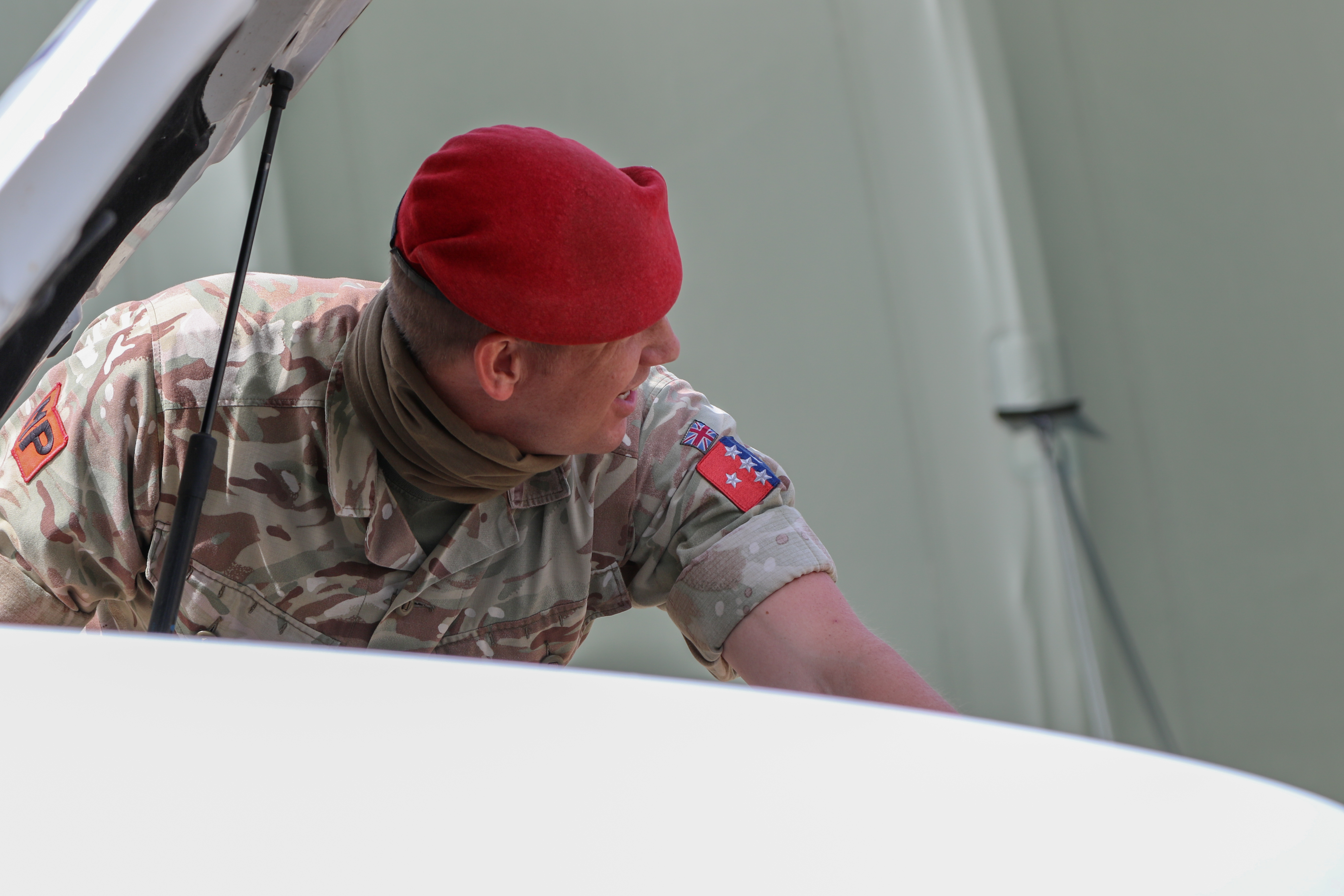 Soldier in camouflage uniform and red beret inspecting a vehicle with its hood open.