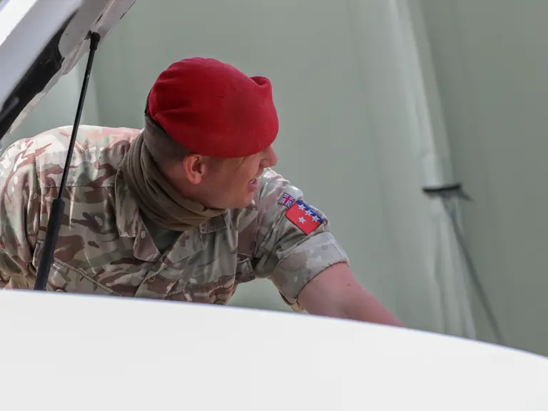 Soldier in camouflage uniform and red beret inspecting a vehicle with its hood open.