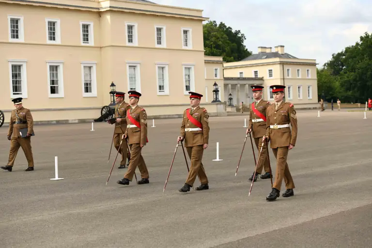 Four British Army soldiers wearing khaki dress uniforms are marching across a parade square using pace sticks. Behind them are two Army personnel watching the team of four.