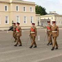 Four British Army soldiers wearing khaki dress uniforms are marching across a parade square using pace sticks. Behind them are two Army personnel watching the team of four.
