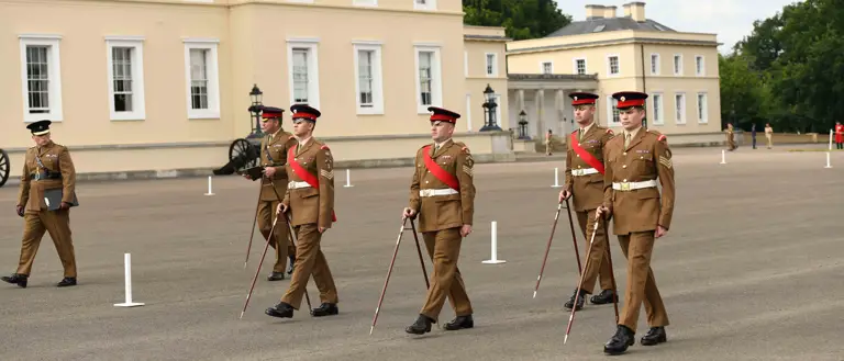 Four British Army soldiers wearing khaki dress uniforms are marching across a parade square using pace sticks. Behind them are two Army personnel watching the team of four.