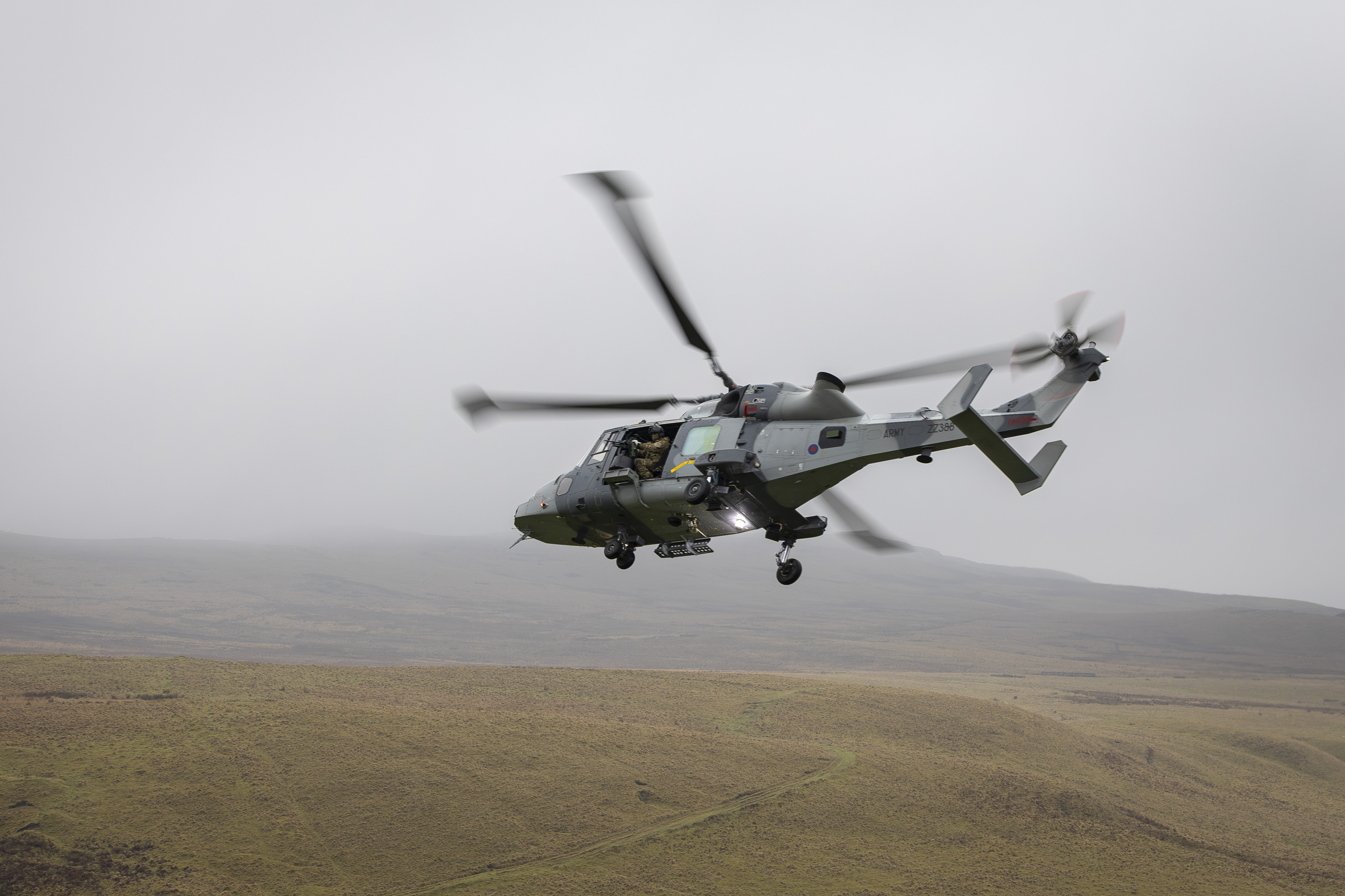 A military helicopter is flying over a vast, grassy landscape with low hills. The sky is overcast.