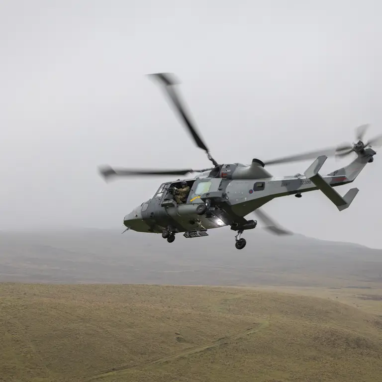 A military helicopter is flying over a vast, grassy landscape with low hills. The sky is overcast.