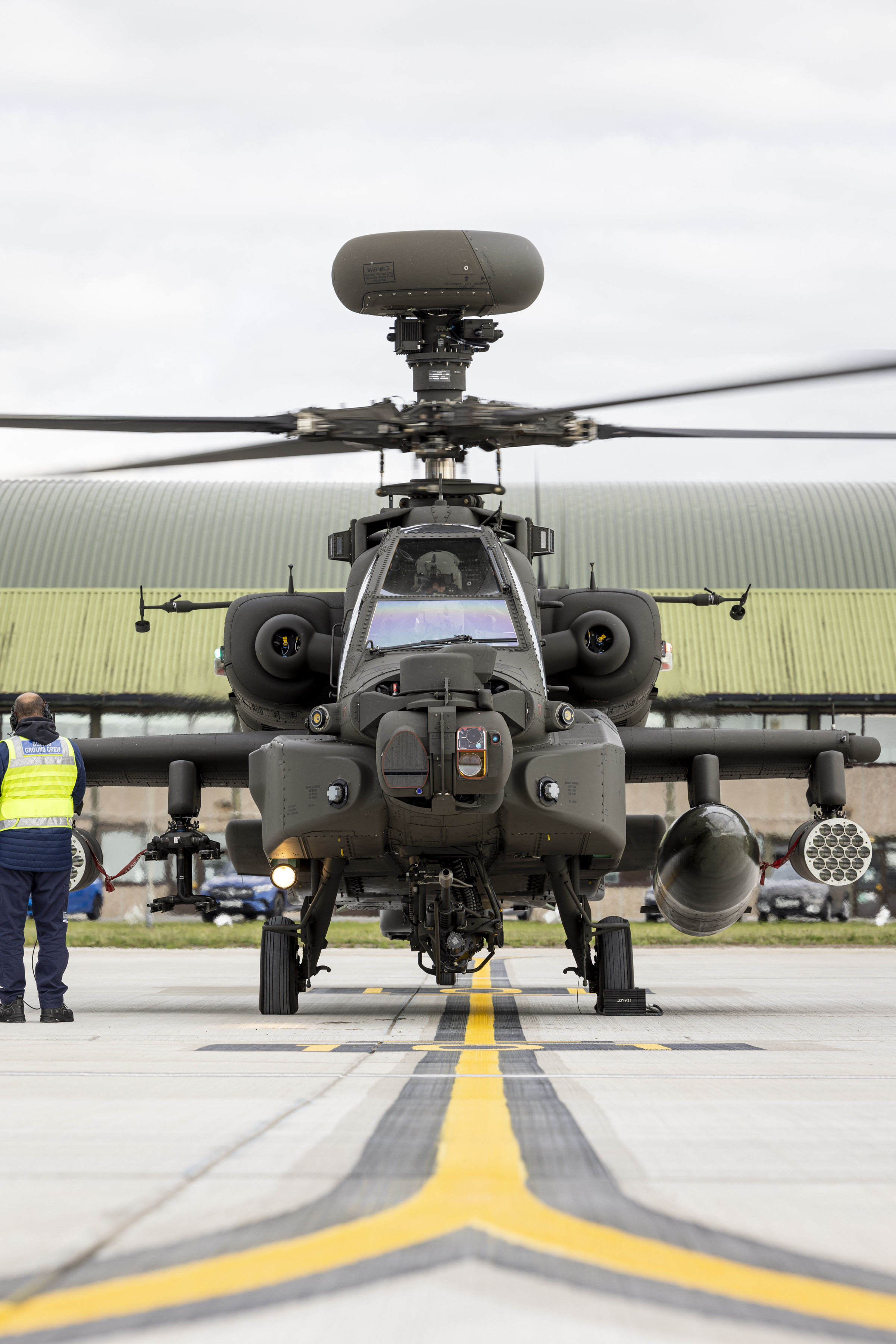 An Apache helicopter on the ground at a military airfield, with a person in a reflective vest nearby.