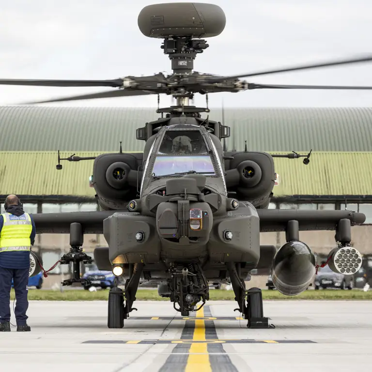 An Apache helicopter on the ground at a military airfield, with a person in a reflective vest nearby.