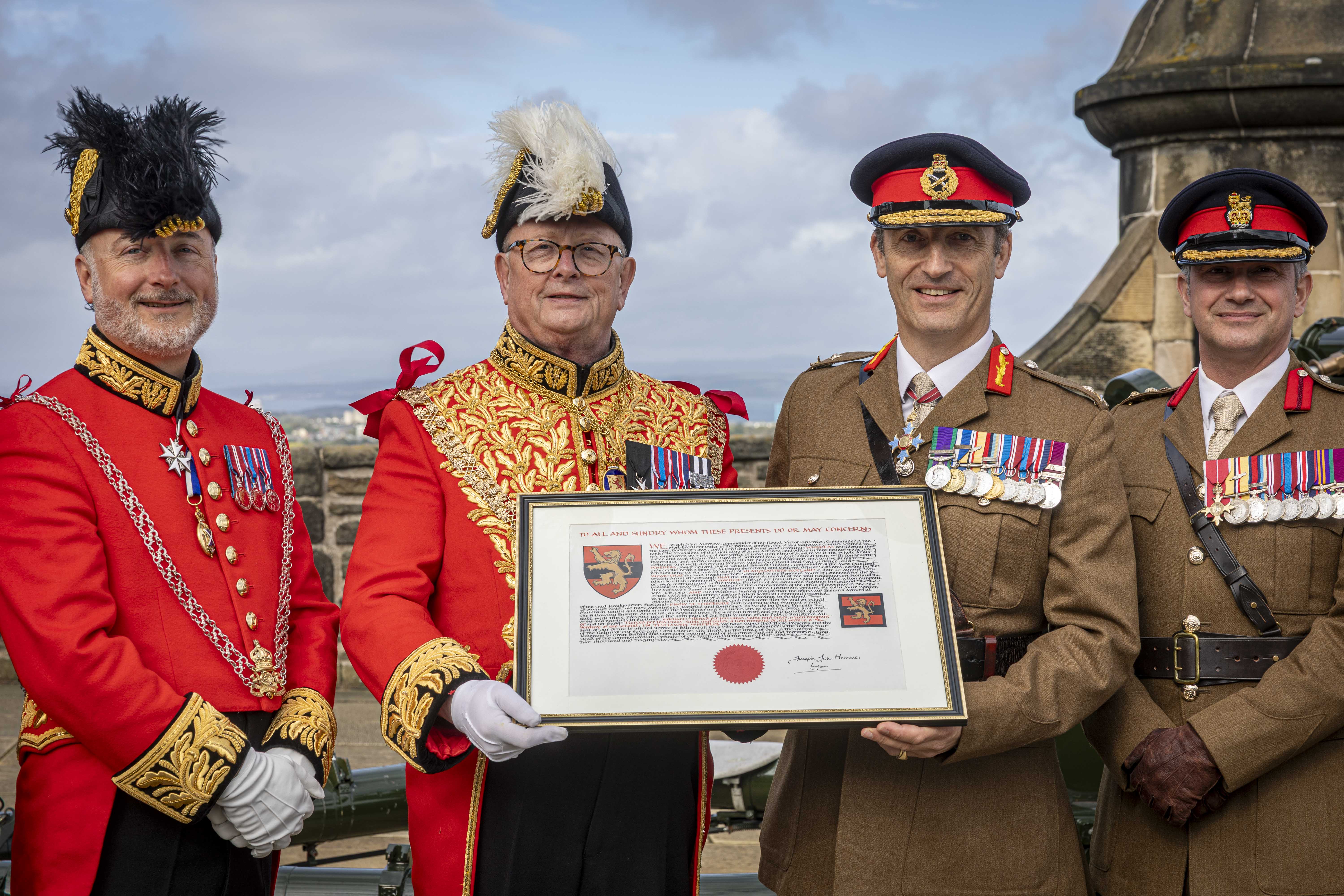 A group of four males in uniform standing front facing side-by-side in a row. Two are wearing bright red ceremonial attire with plumed hats, and the other two are in khaki military uniforms with black hats peaked caps. The middle two are holding a framed document. All four are displaying medals on their chest and are standing in the one o'clock gun salute area of Edinburgh Castle. 