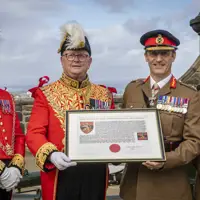 A group of four males in uniform standing front facing side-by-side in a row. Two are wearing bright red ceremonial attire with plumed hats, and the other two are in khaki military uniforms with black hats peaked caps. The middle two are holding a framed document. All four are displaying medals on their chest and are standing in the one o'clock gun salute area of Edinburgh Castle.