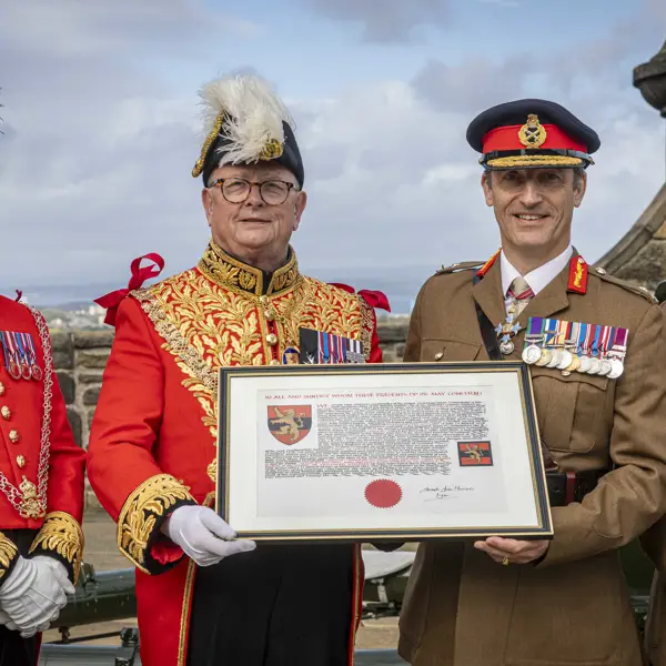 A group of four males in uniform standing front facing side-by-side in a row. Two are wearing bright red ceremonial attire with plumed hats, and the other two are in khaki military uniforms with black hats peaked caps. The middle two are holding a framed document. All four are displaying medals on their chest and are standing in the one o'clock gun salute area of Edinburgh Castle.