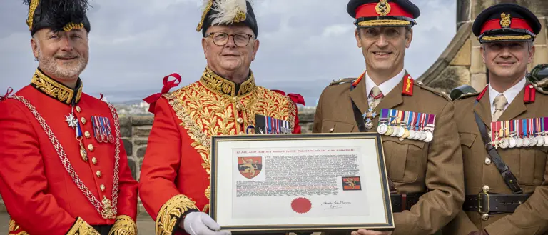 A group of four males in uniform standing front facing side-by-side in a row. Two are wearing bright red ceremonial attire with plumed hats, and the other two are in khaki military uniforms with black hats peaked caps. The middle two are holding a framed document. All four are displaying medals on their chest and are standing in the one o'clock gun salute area of Edinburgh Castle.