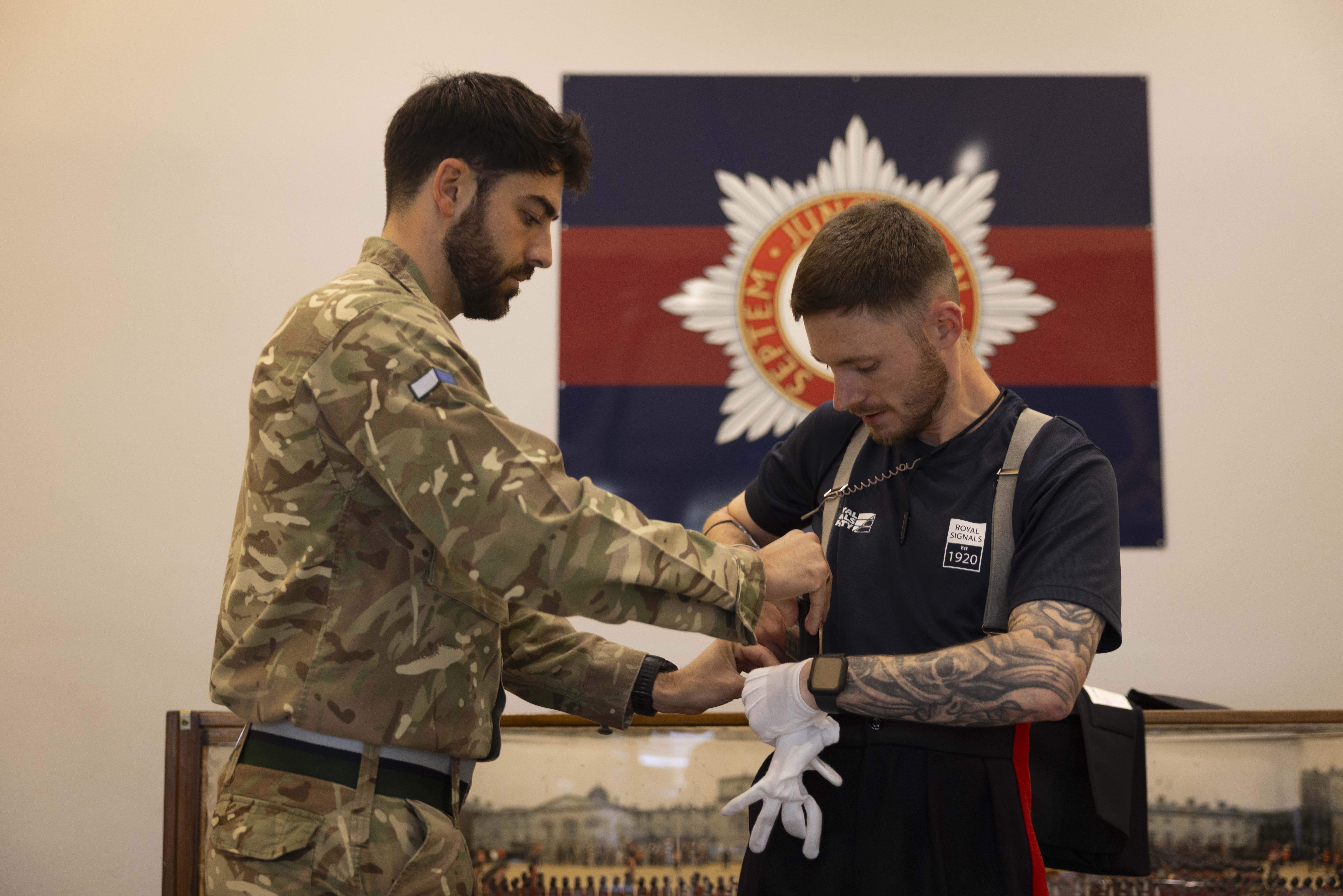 A soldier in camouflage assists a man wearing athletic clothing with his uniform and gloves. They stand in front of a military emblem on the wall.