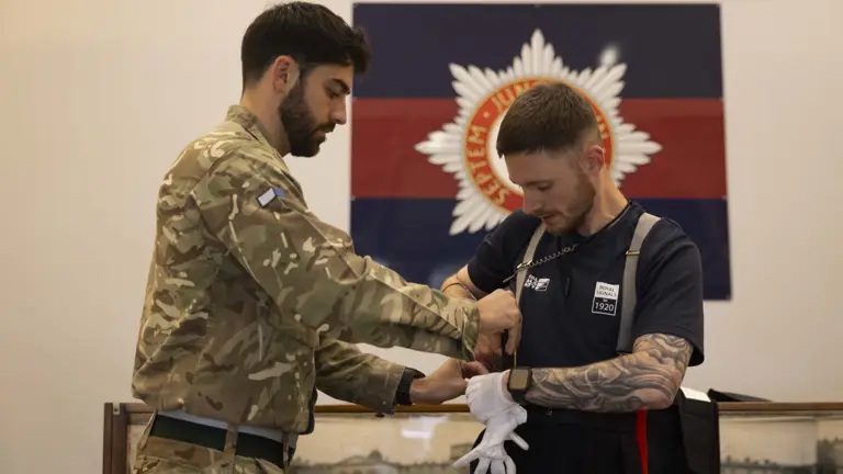 A soldier in camouflage assists a man wearing athletic clothing with his uniform and gloves. They stand in front of a military emblem on the wall.