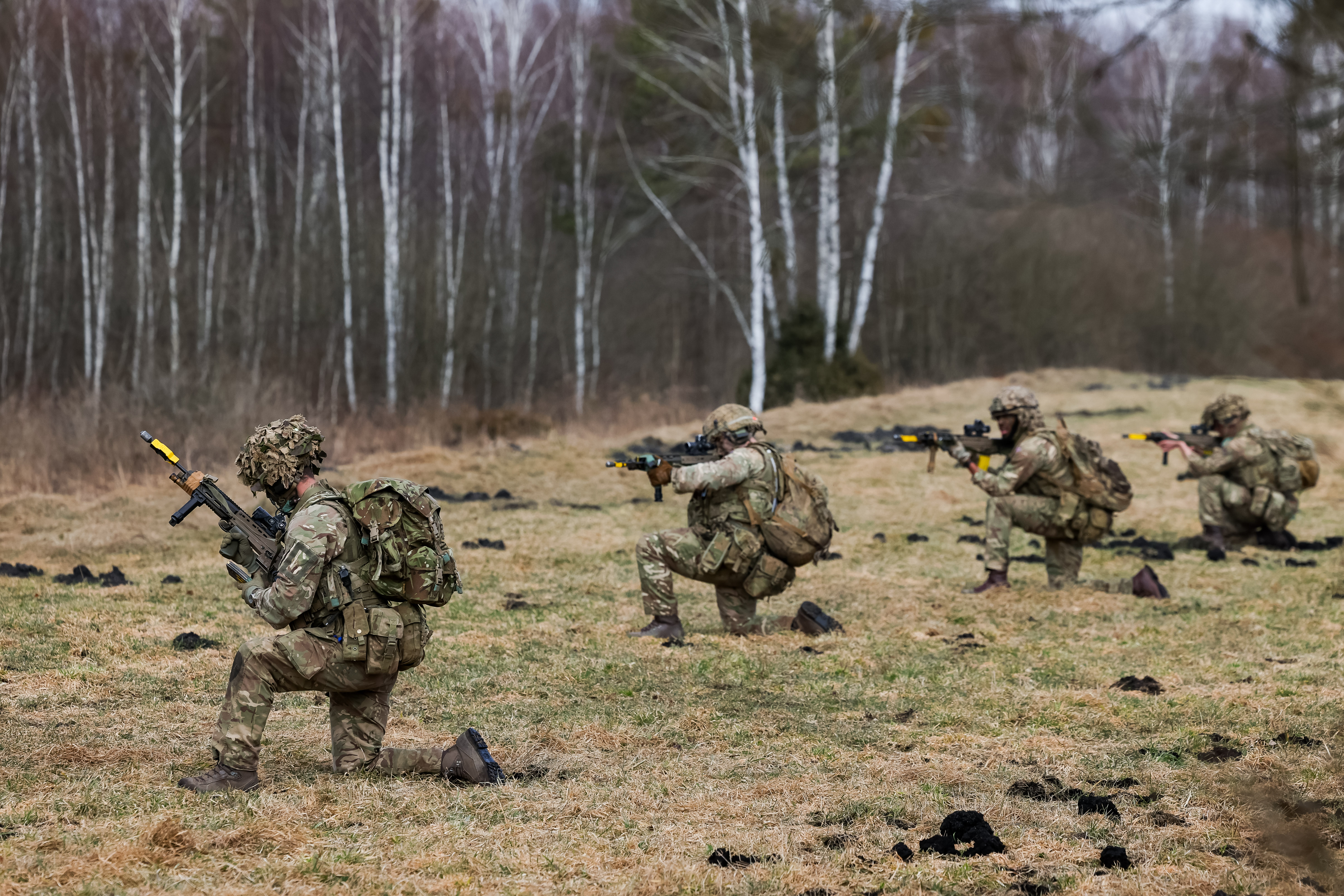 Four soldiers in camouflage uniforms kneel while taking aim with their rifles.