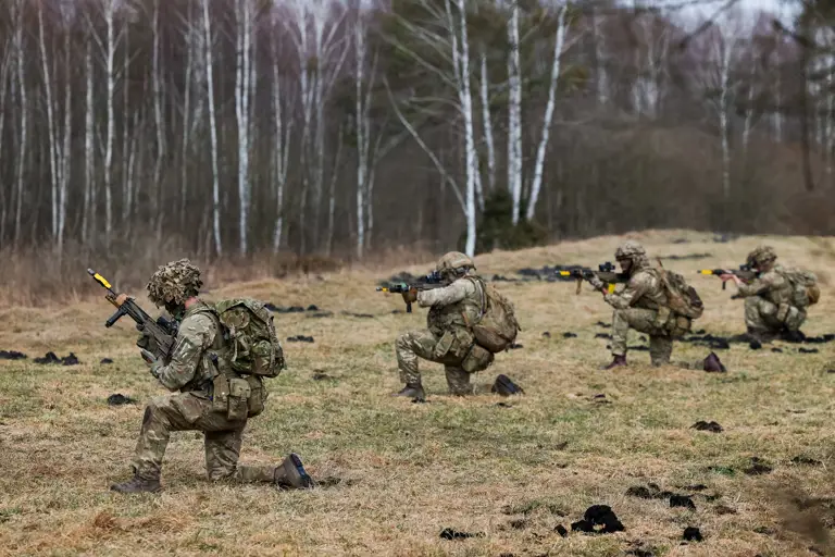 Four soldiers in camouflage uniforms kneel while taking aim with their rifles.