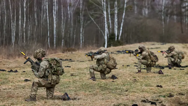 Four soldiers in camouflage uniforms kneel while taking aim with their rifles.
