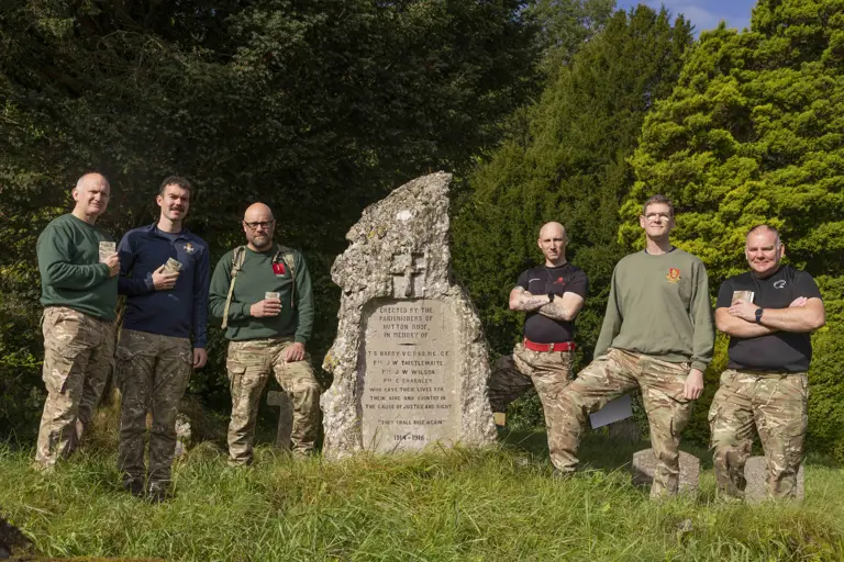 Six soldiers wearing camouflage trousers and a jumper, stand and pose for a photo at the end of their charity walk but a stone memorial plaque.