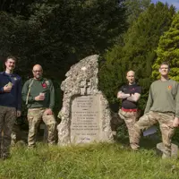 Six soldiers wearing camouflage trousers and a jumper, stand and pose for a photo at the end of their charity walk but a stone memorial plaque.