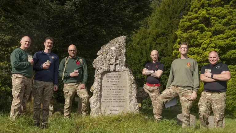 Six soldiers wearing camouflage trousers and a jumper, stand and pose for a photo at the end of their charity walk but a stone memorial plaque.