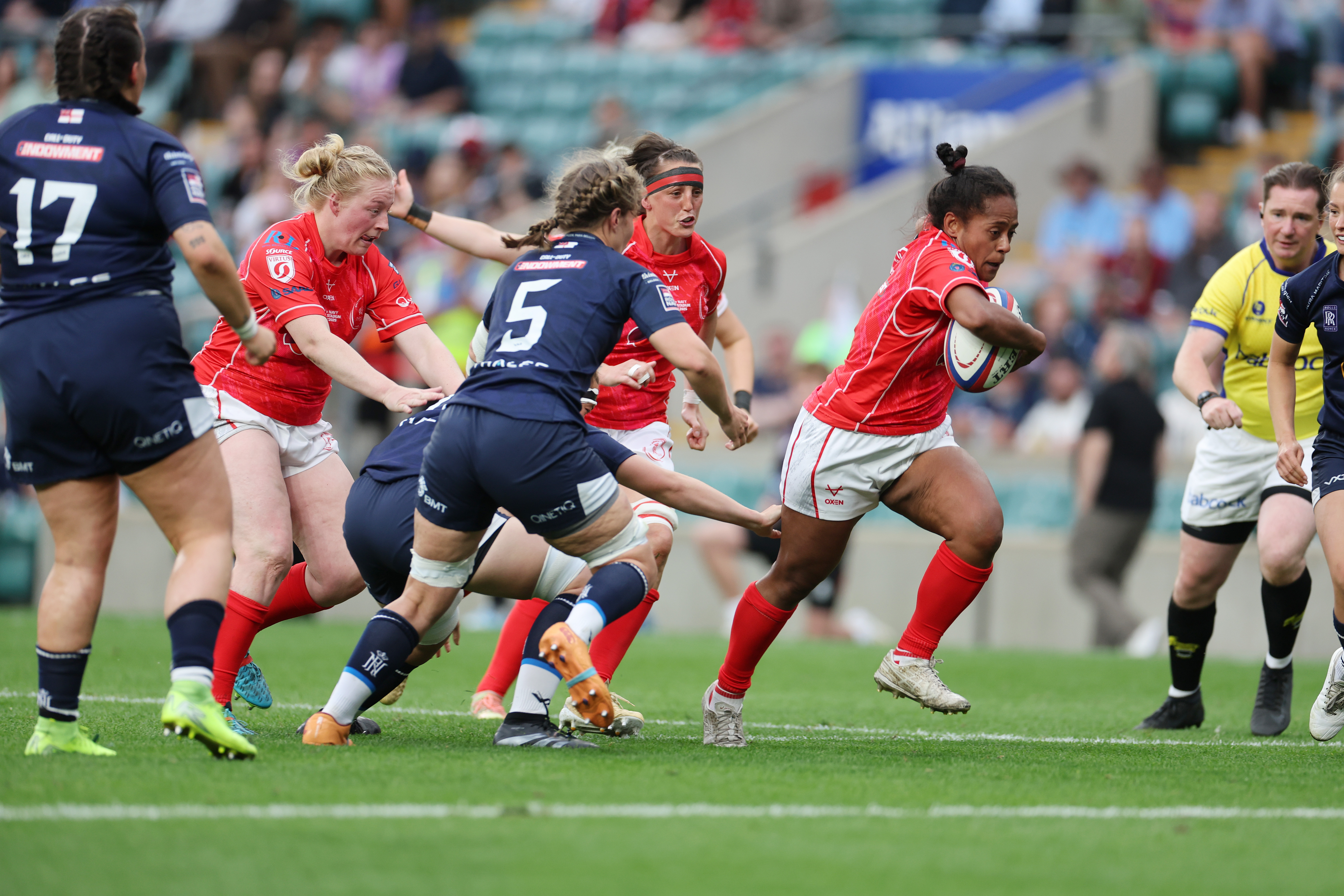Female rugby player in red jersey runs with the ball, evading opponents in blue. Teammates and referee watch intensely. Energetic and competitive scene.