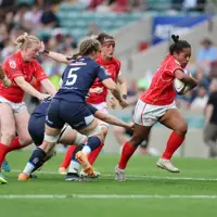 Female rugby player in red jersey runs with the ball, evading opponents in blue. Teammates and referee watch intensely. Energetic and competitive scene.