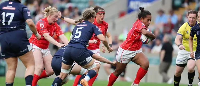 Female rugby player in red jersey runs with the ball, evading opponents in blue. Teammates and referee watch intensely. Energetic and competitive scene.