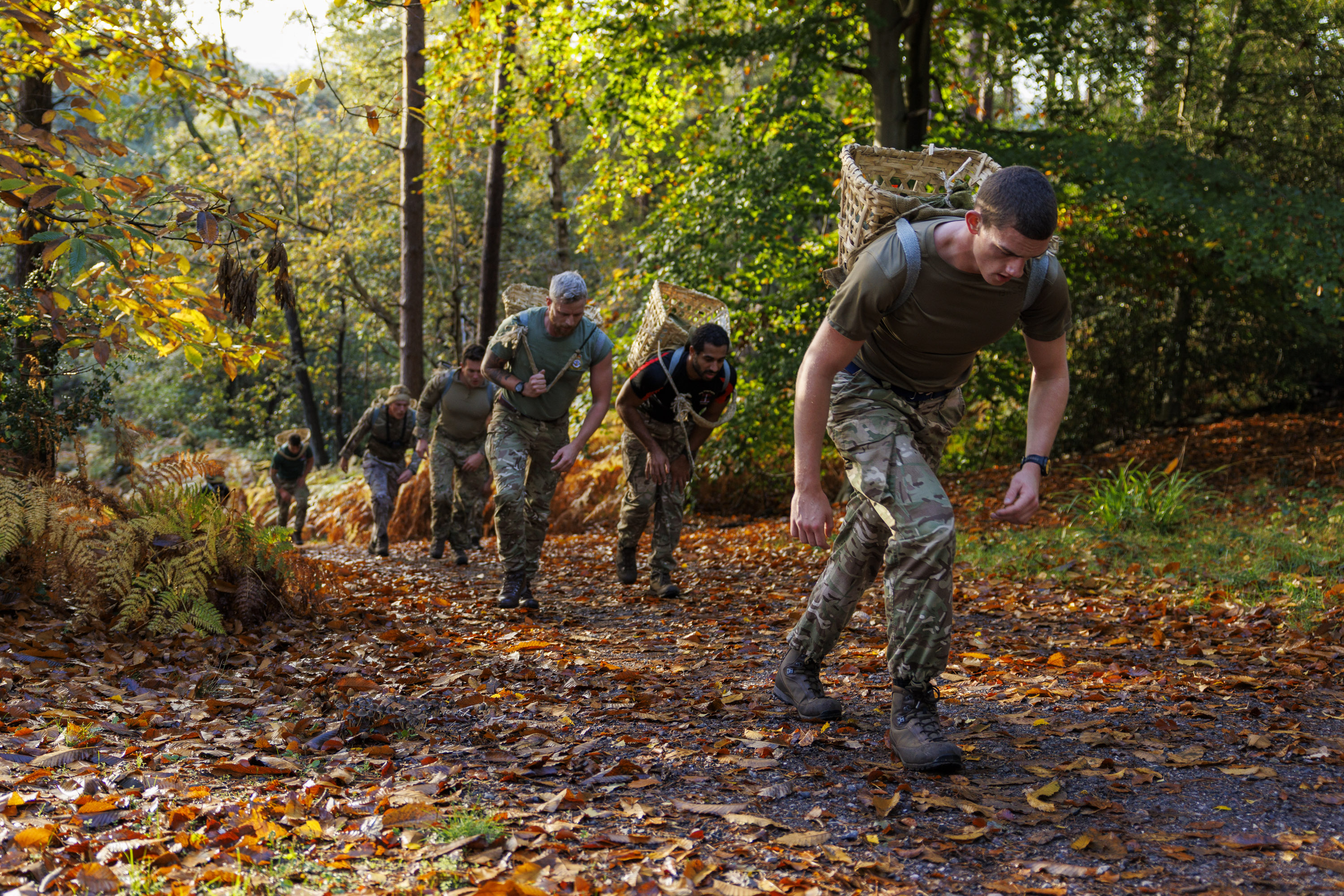 Group of soldiers in camouflage uniforms running uphill through a forest trail covered with autumn leaves.