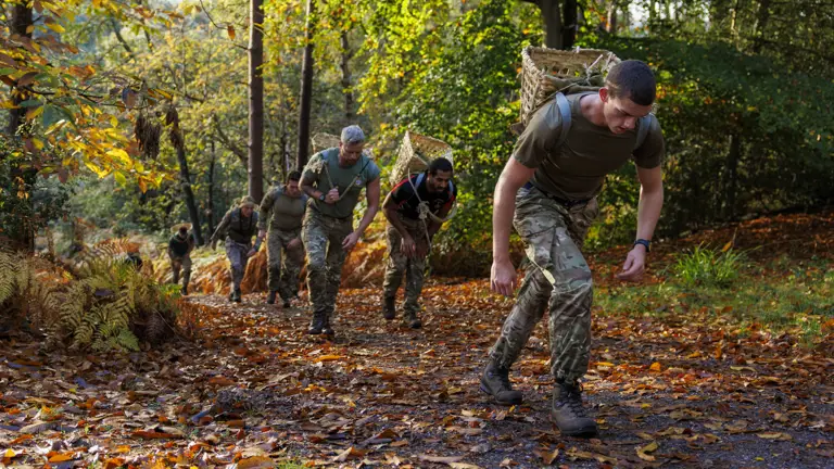 Group of soldiers in camouflage uniforms running uphill through a forest trail covered with autumn leaves.