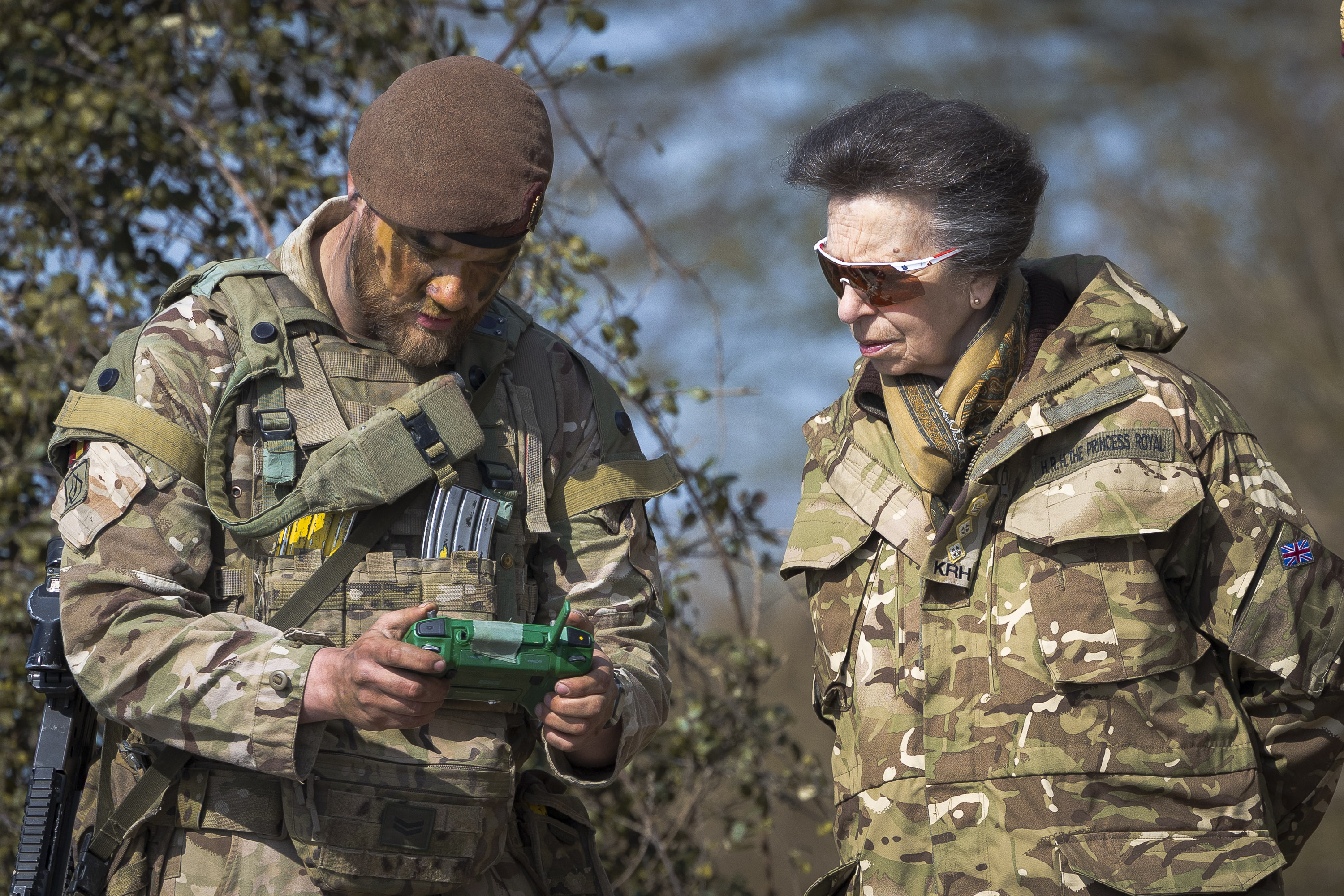 A person in military gear is using a device while another, in similar camouflage attire, observes. 