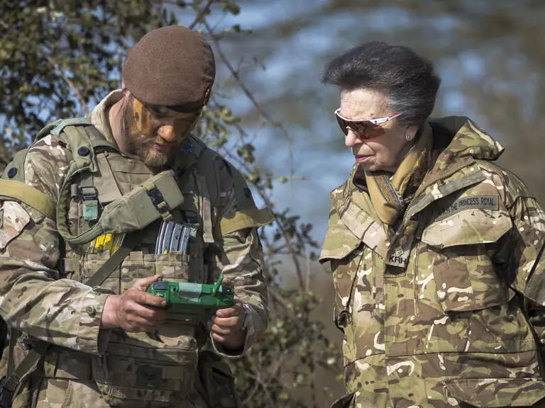 A person in military gear is using a device while another, in similar camouflage attire, observes.