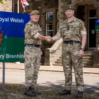 Two men in camouflage uniforms stand in front of a building shaking hands.