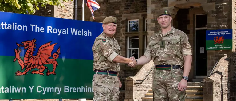 Two men in camouflage uniforms stand in front of a building shaking hands.