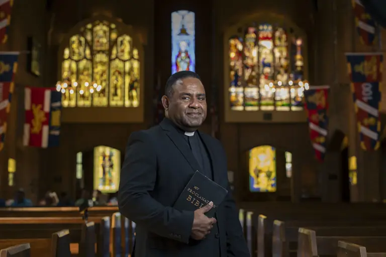 Clergyman in black suit holding a Bible inside a church with stained glass windows and colorful banners.