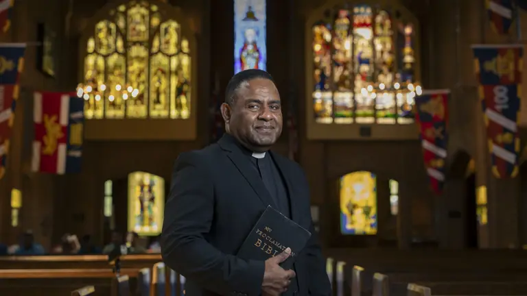 Clergyman in black suit holding a Bible inside a church with stained glass windows and colorful banners.