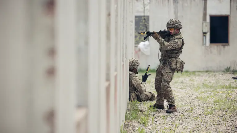 A soldier wearing camouflage uniform holds a rifle up to a window. There is another soldier sat at his feet.