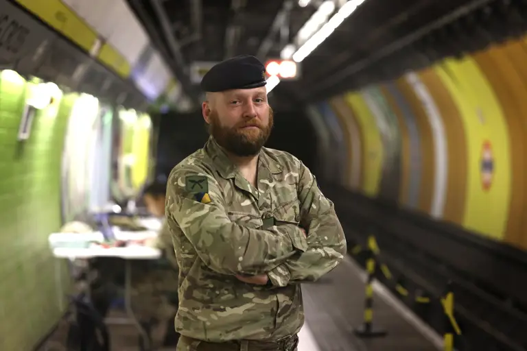 An individual in camouflage military uniform and beret stands confidently in a London Underground station.