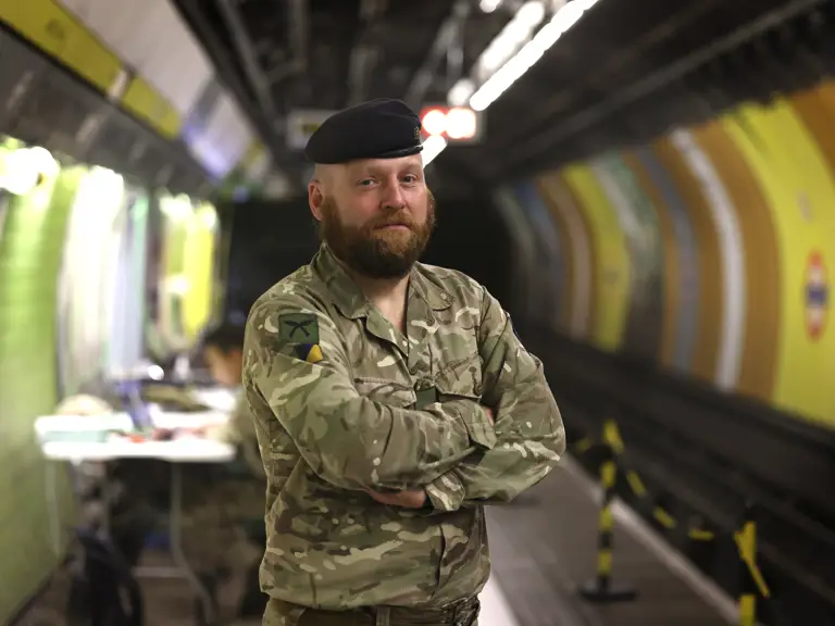 An individual in camouflage military uniform and beret stands confidently in a London Underground station.