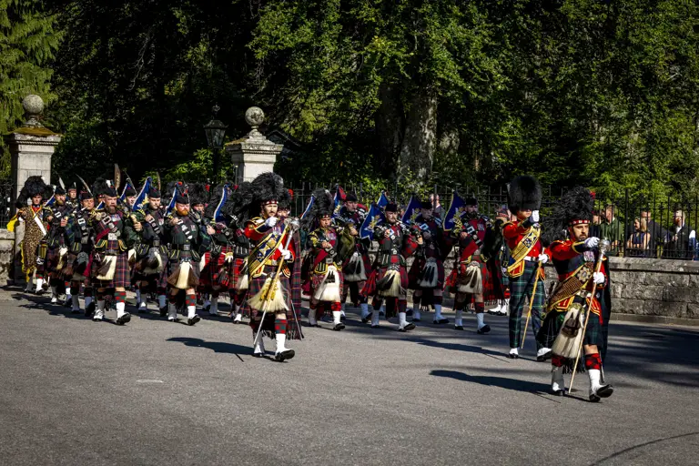 A Scottish pipe band in traditional tartan kilts march in formation on a sunny day.
