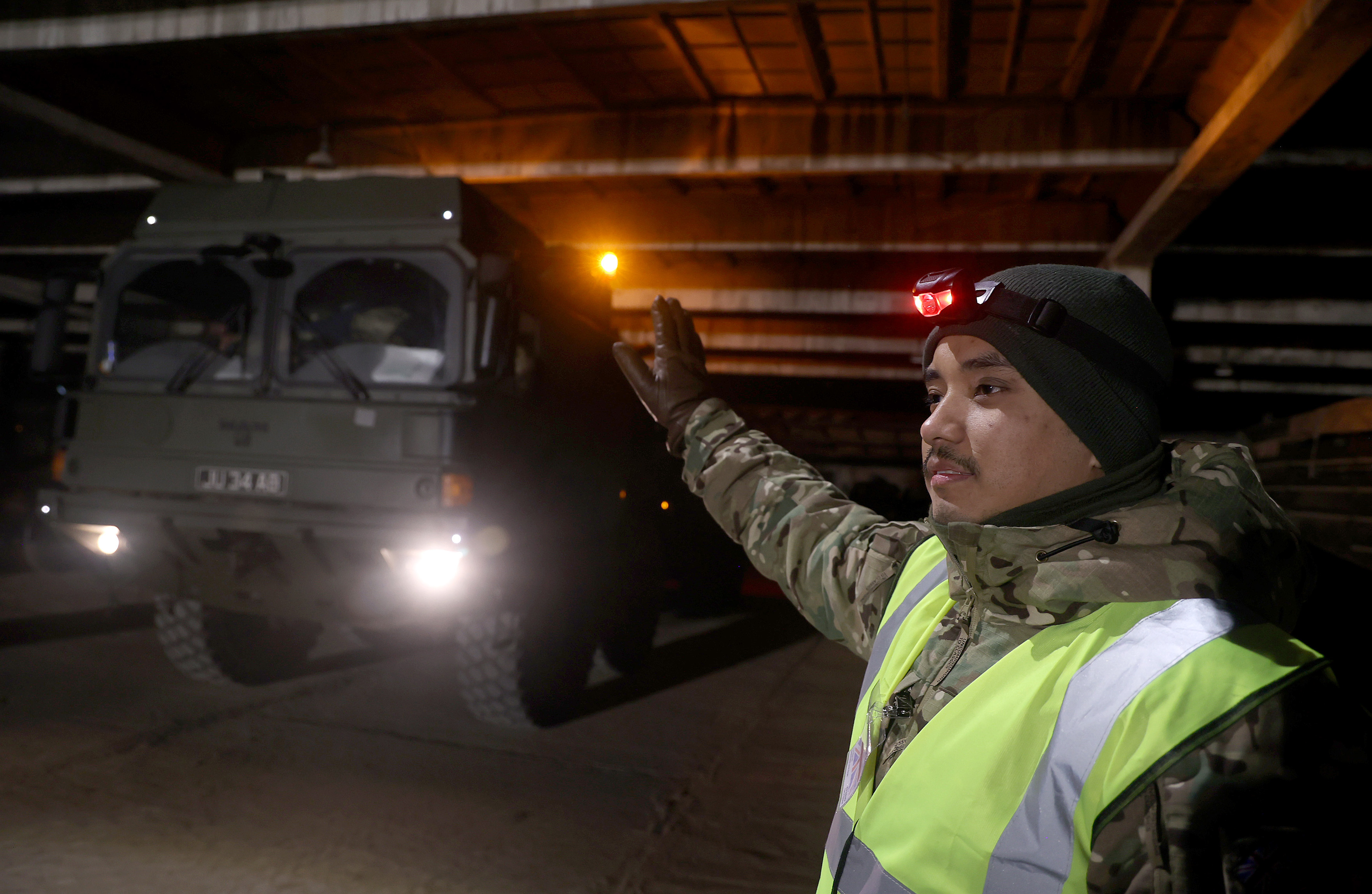 Large army transport vehicle being guided by a soldier in camouflage uniform at night.