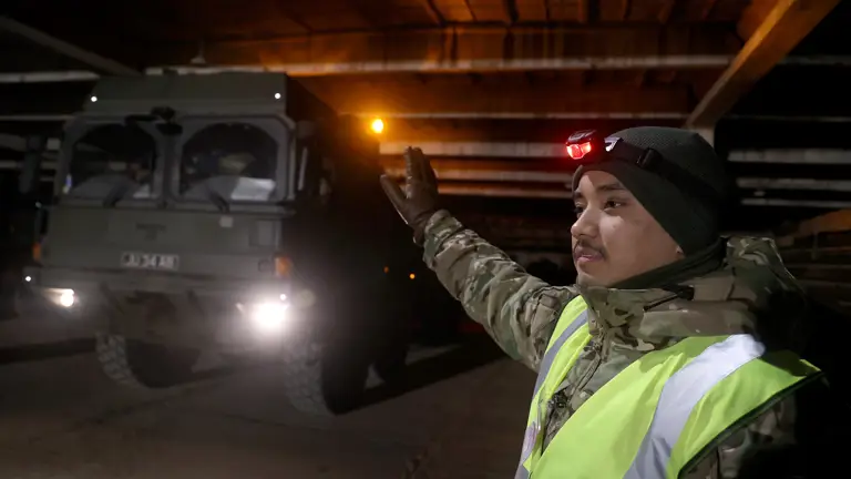 Large army transport vehicle being guided by a soldier in camouflage uniform at night.
