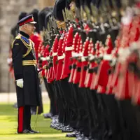 His Majesty The King smiles at soldiers wearing red tunics and bearskin hats, the soldiers have their weapons raised.