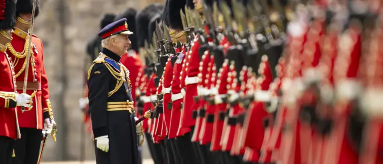 His Majesty The King smiles at soldiers wearing red tunics and bearskin hats, the soldiers have their weapons raised.