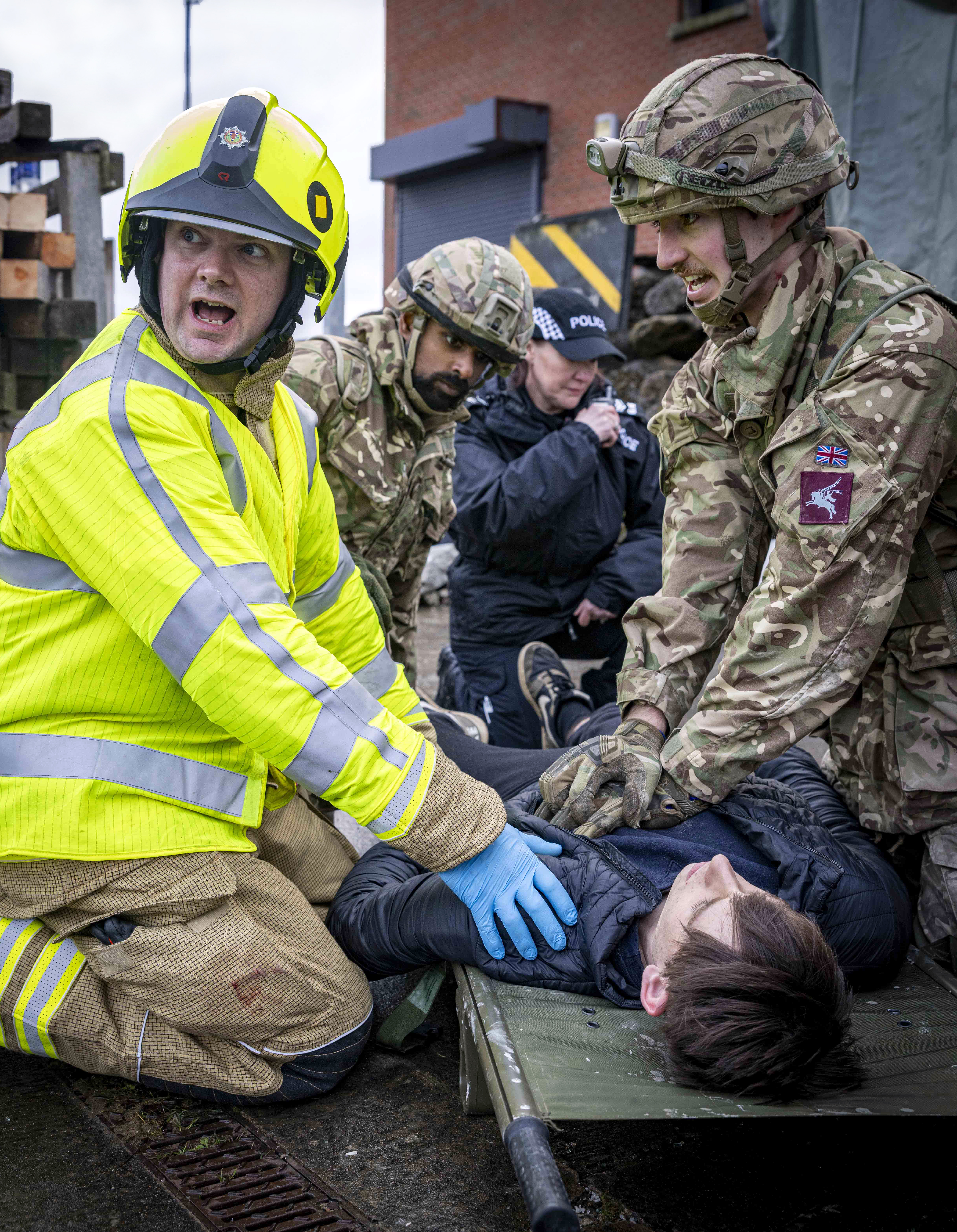 Emergency responder in high-visibility gear and soldiers attending to a person lying on a stretcher during a rescue drill.