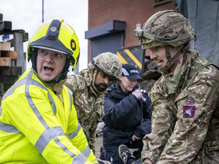 Emergency responder in high-visibility gear and soldiers attending to a person lying on a stretcher during a rescue drill.