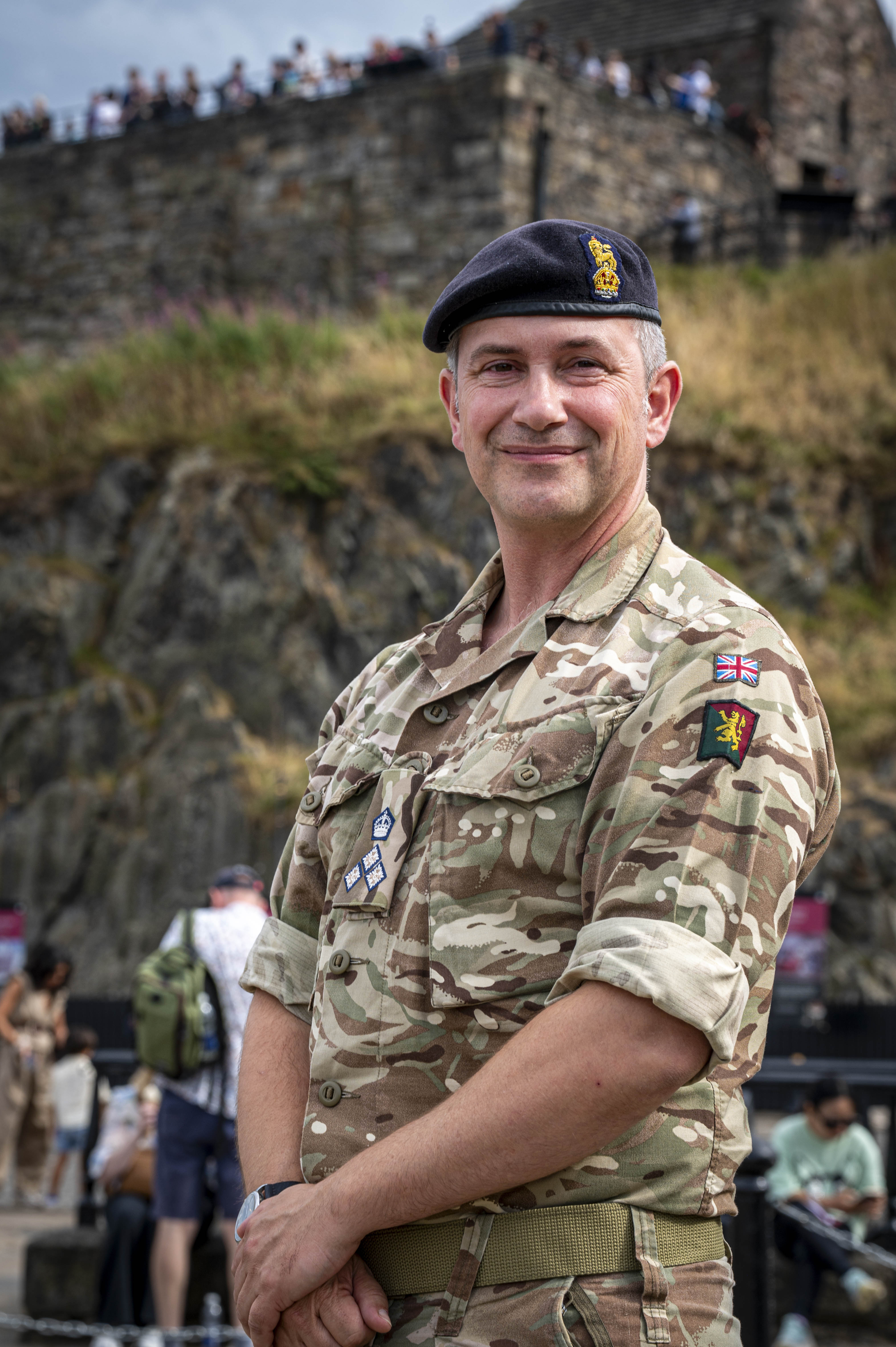 One individual in military uniform, wearing a beret with insignia, standing confidently with arms crossed. They are outdoors with an architectural structure visible in the background.